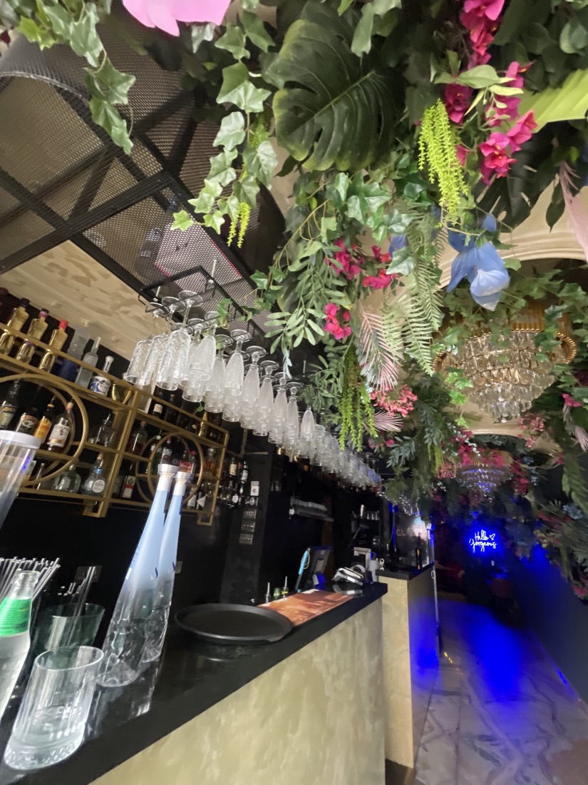 The Globe Lounge bar framed by hanging plants and pink flowers, gold shelving, a crystal chandelier and a blue neon 'Hello Gorgeous' sign
