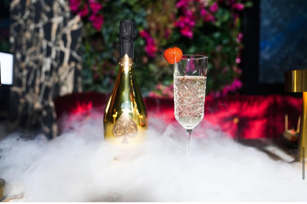 A gold champagne bottle and a crystal-cut flute wreathed in dry-ice smoke, against magenta flowers and red velvet seating