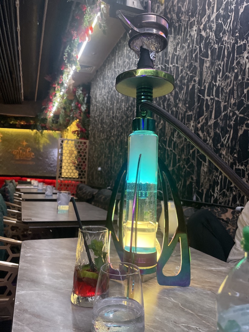 An iridescent shisha pipe glowing green and gold on a marble table, with a fresh mocktail beside it